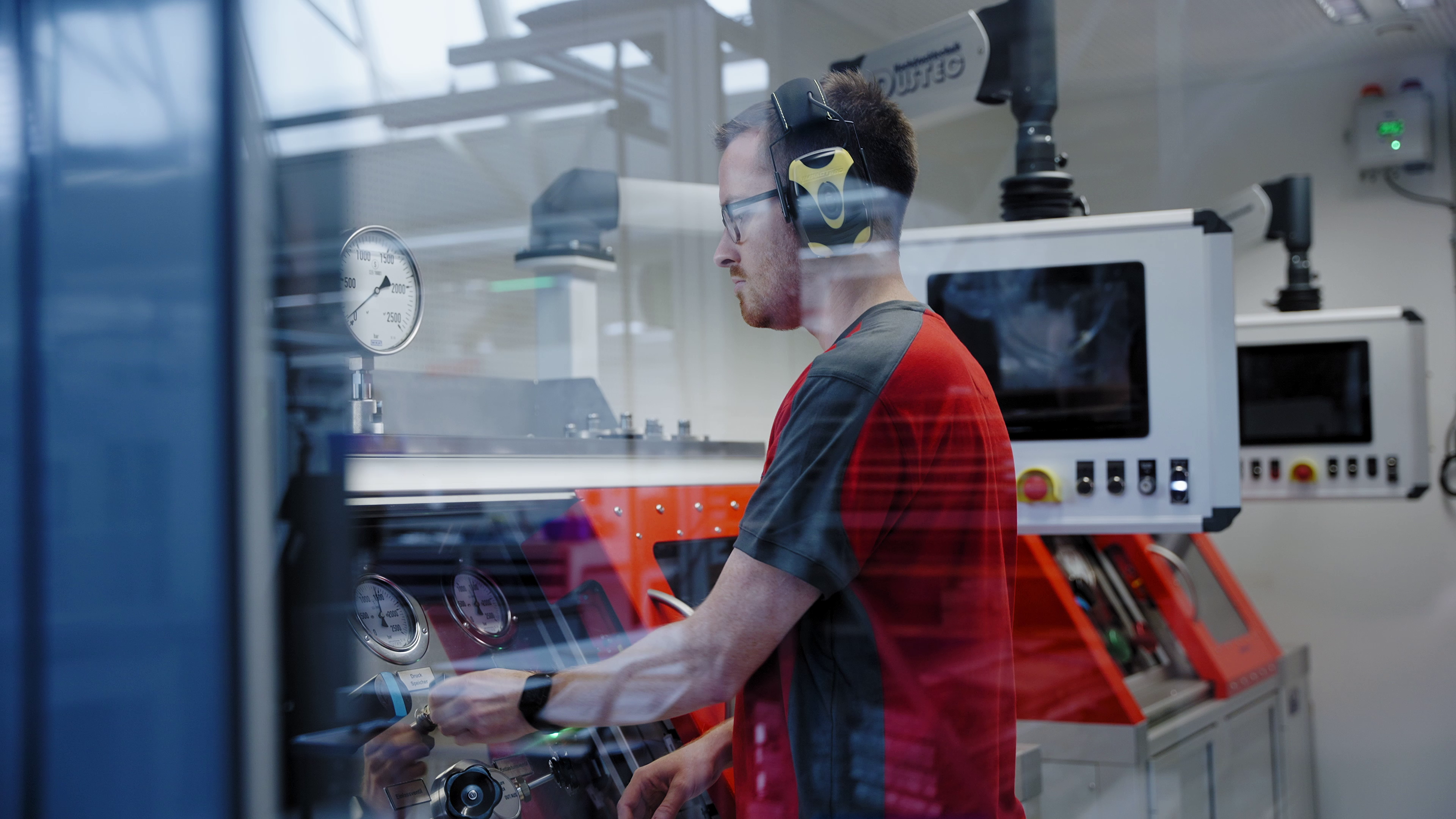 Ein Mann in roter Arbeitskleidung mit Ohrschützern bedient hinter einem Glas eine Maschine